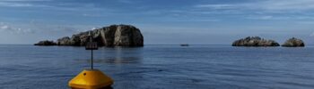 Κίτρινος φωτοσημαντήρας στο Καταδυτικό Πάρκο των Φούρνων, με τις βραχονησίδες στις Λεμονόπετρες στο βάθος.