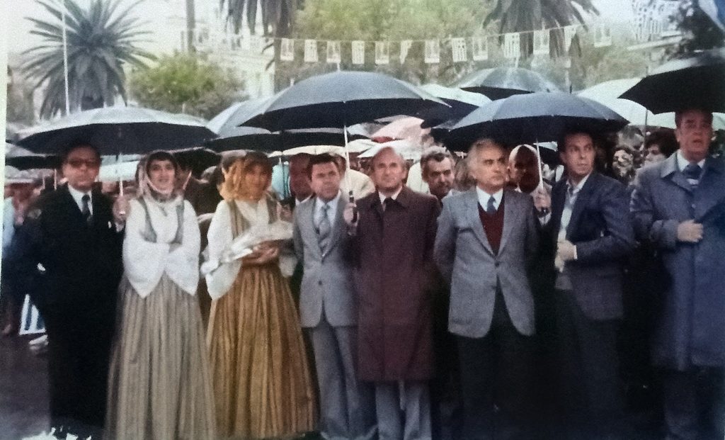 Μέλη του Δημοτικού Συμβουλίου Σαμίων περιμένουν με ομπρέλες τον Ανδρέα Παπανδρέου στην πλατεία Πυθαγόρα το 1983.
