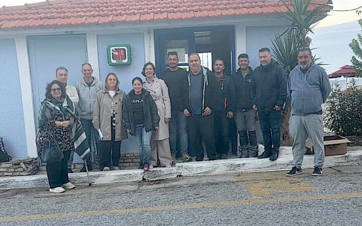 Ομαδική φωτογραφία από την παράδοση απινιδωτή για το Ηραίο, έξω από κτίριο με επιτοίχια θήκη απινιδωτή.