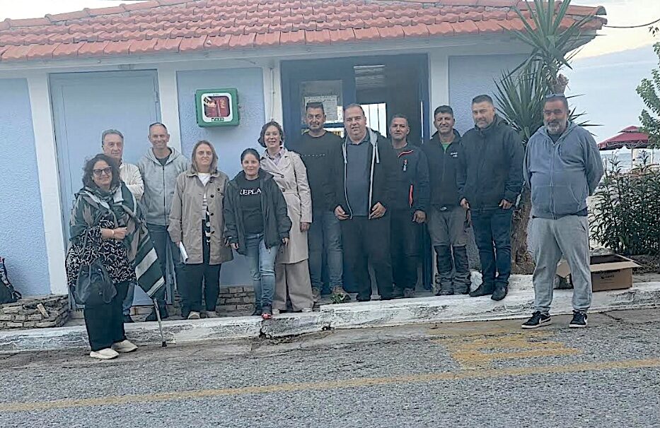 Ομαδική φωτογραφία από την παράδοση απινιδωτή για το Ηραίο, έξω από κτίριο με επιτοίχια θήκη απινιδωτή.